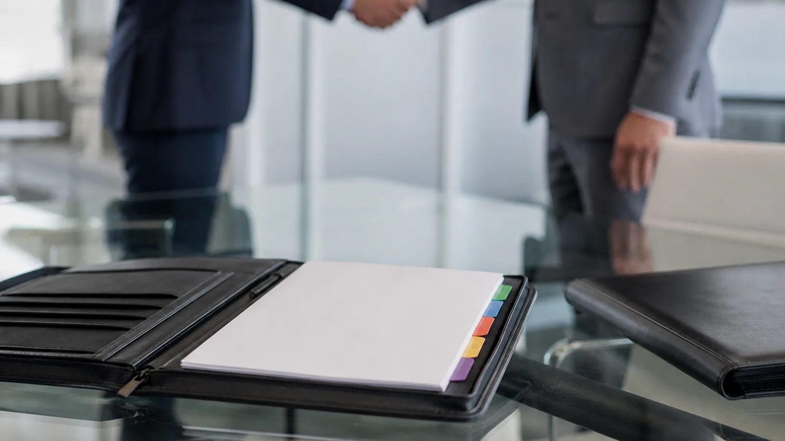 A leather portfolio open on a conference table showing blank tabbed proposal sections, a handshake in soft focus background.
