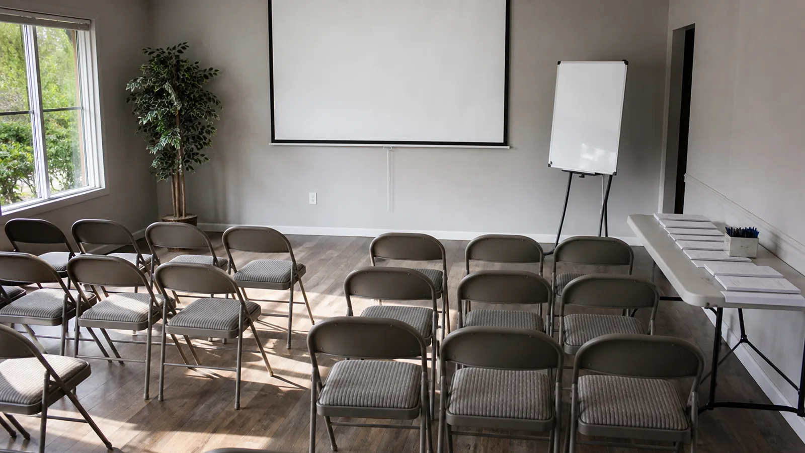 A community classroom with rows of folding chairs, a blank projected screen at the front, and a stack of workshop handouts face-down on a side table.