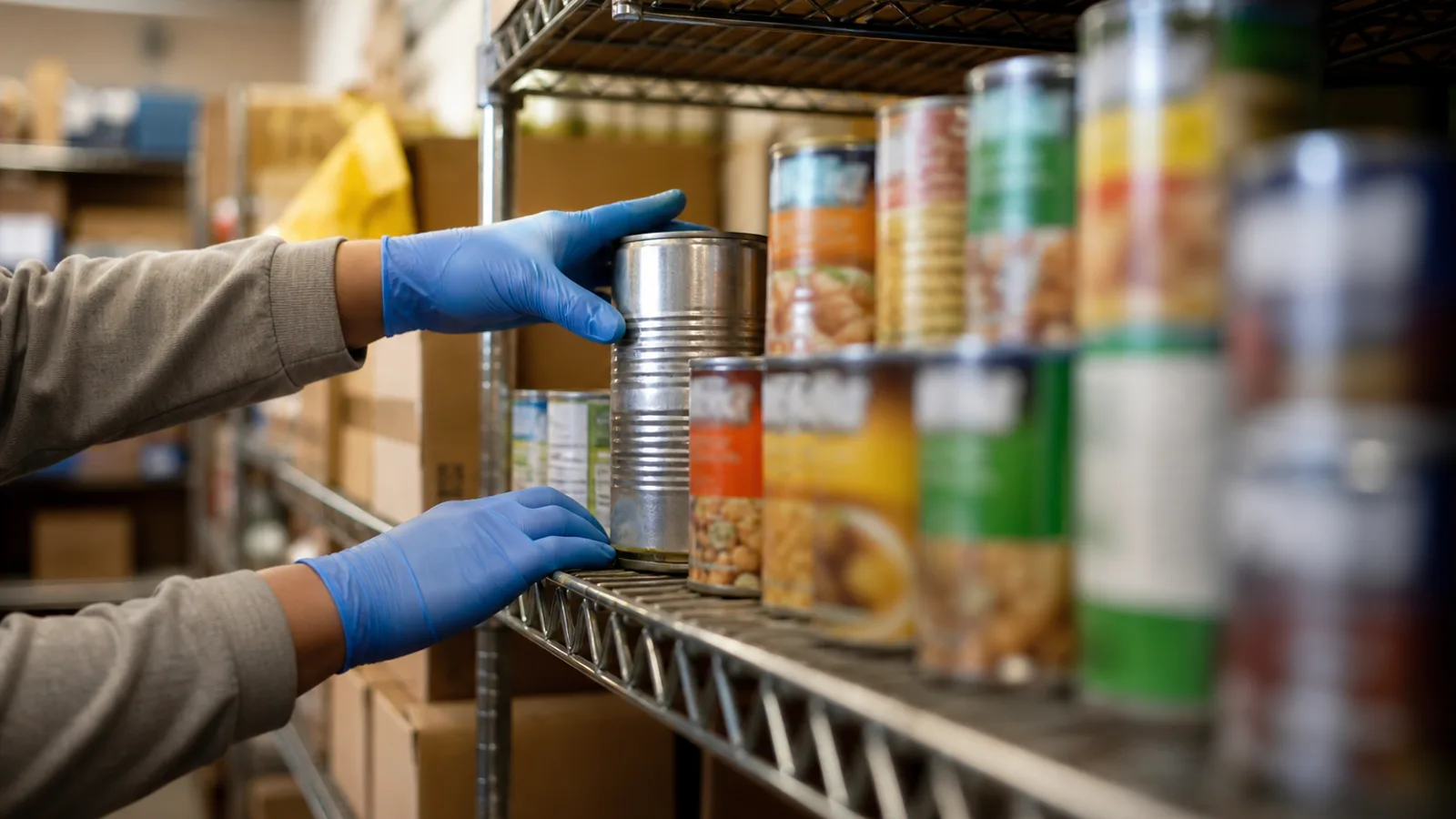 A volunteer's hands carefully placing a labeled food item on a community food bank shelf, organized boxes visible in the background.